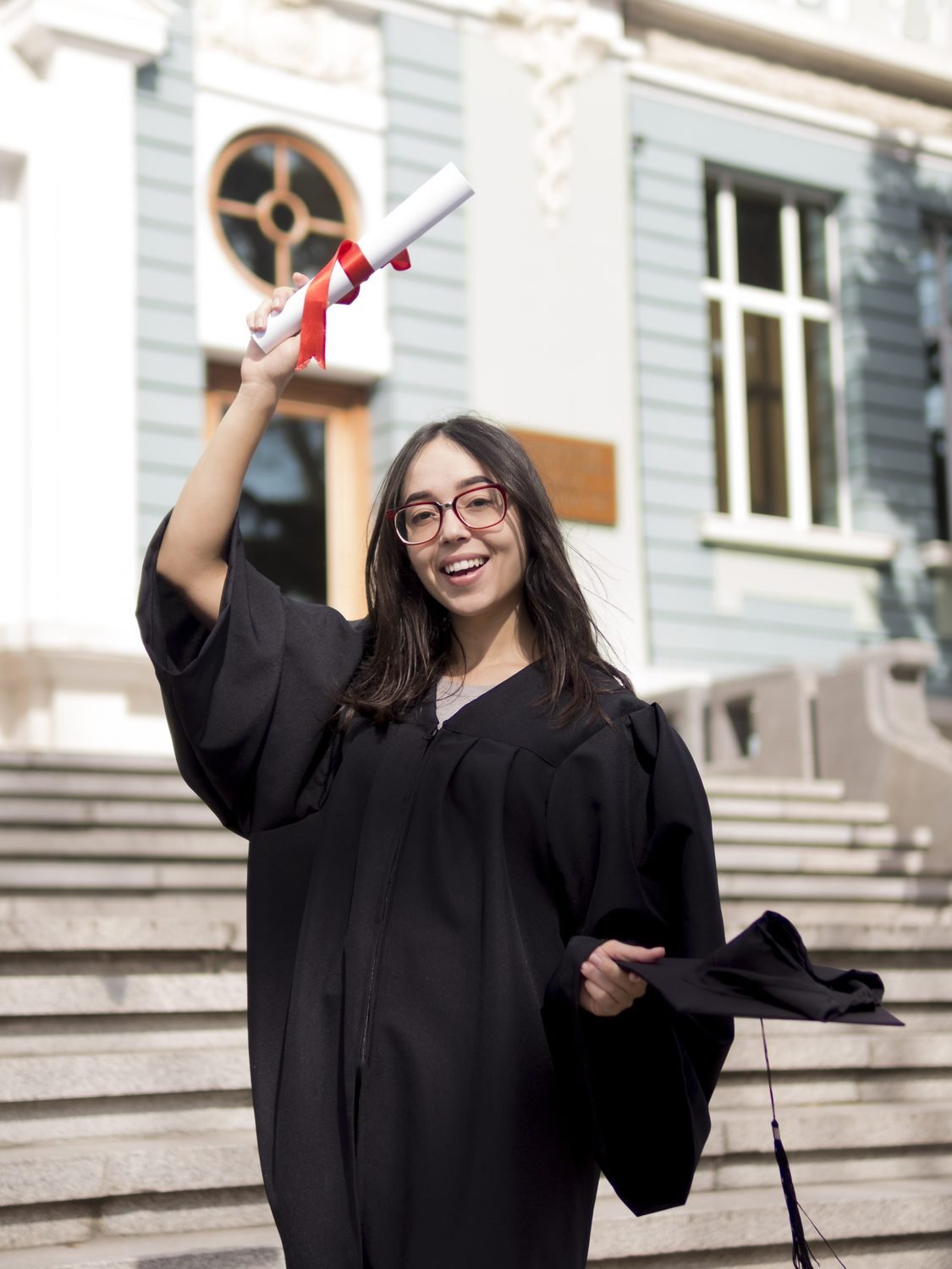 Joven graduada sonriendo y levantando su diploma frente a la universidad